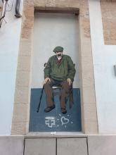 Street art: Older man sitting on a chair, a cane next to him and wearing a face mask.